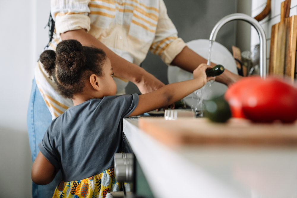 Child washing vegetables at the kitchen sink alongside a parent, practicing daily living skills at home.