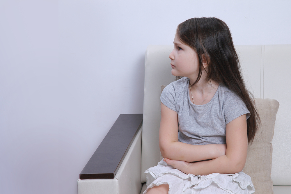 A girl with long brown hair and arms crossed sitting on a chair wearing a grey T-shirt and white skirt.