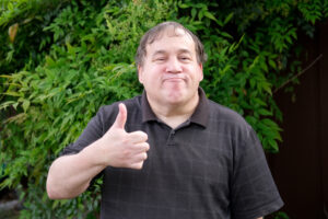 An older autistic man smiling at the camera and giving a thumbs up.