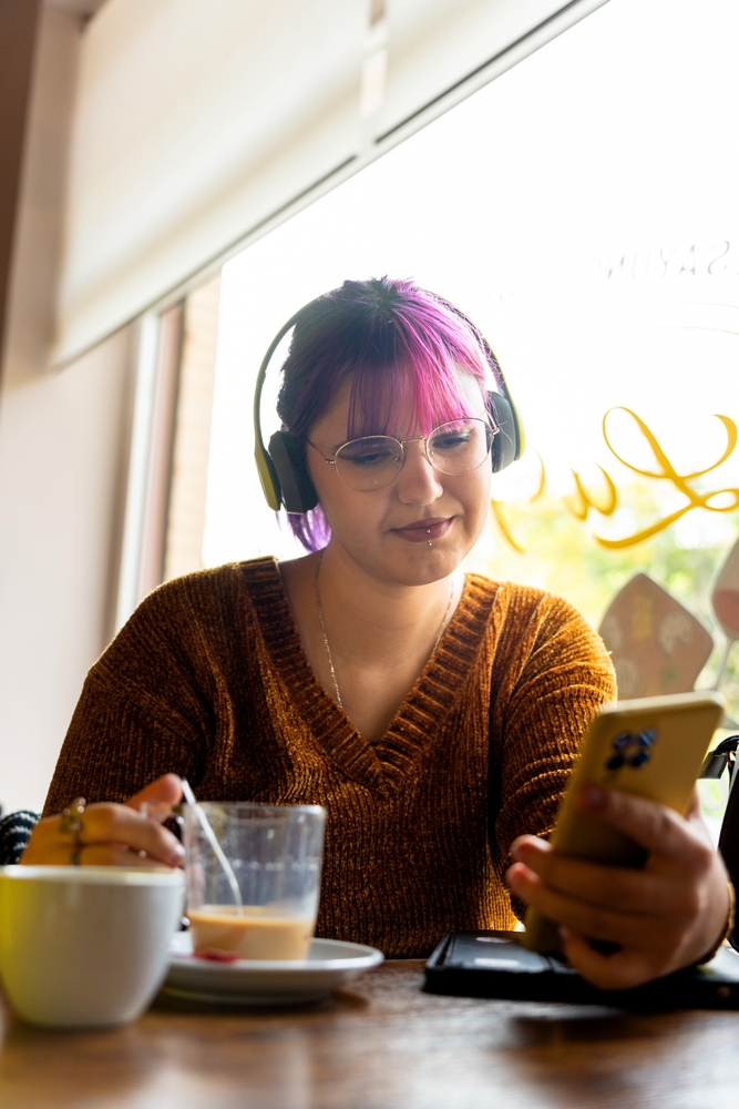 A young woman with glasses, an orange sweater, and pink hair, listening to music coming from her phone.