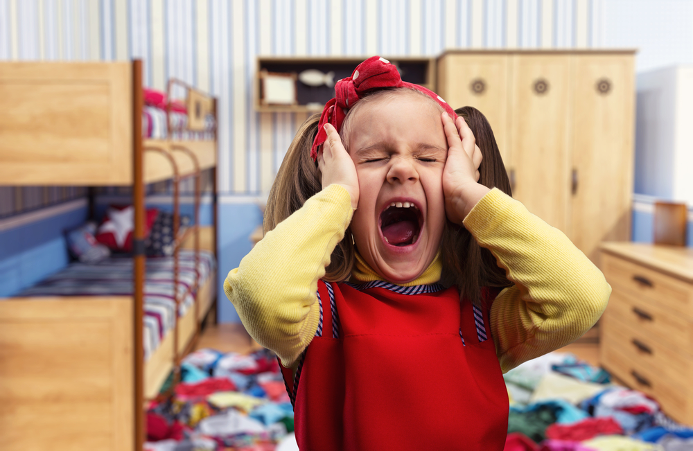 Brown-haired girl wearing a yellow sweater and red dress covering her ears, with her eyes closed, screaming at the top of her lungs.
