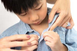 A young Asian boy getting help from an adult to do up the buttons on his dress shirt, while he tries to do it himself.