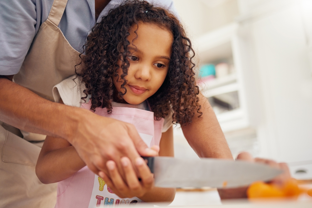 A young black girl wearing an apron, with some help from her dad, as she learns to use a knife to cut in the kitchen.