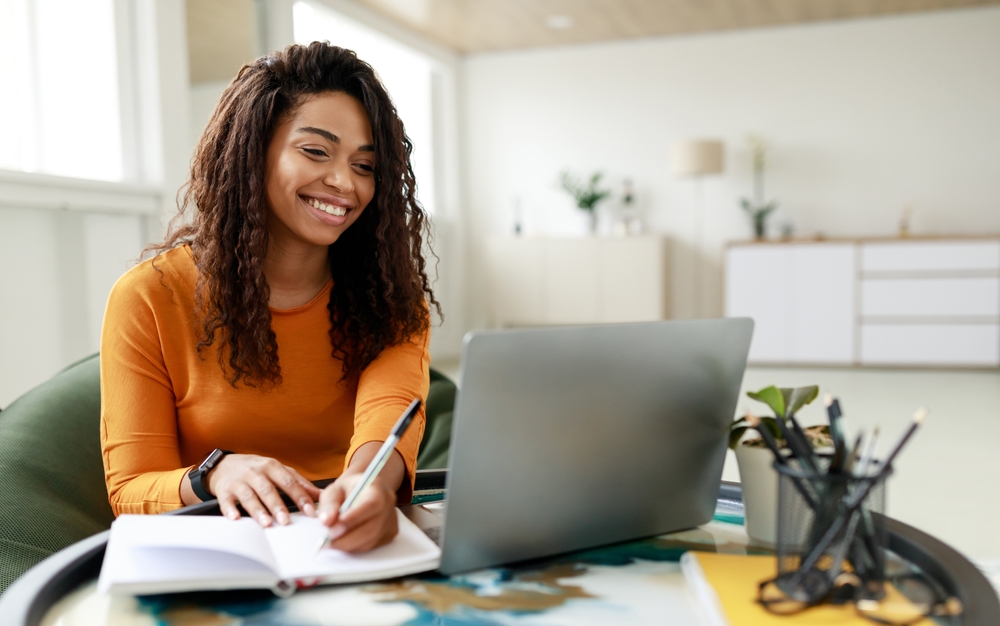 A young black woman wearing an orange long-sleeve shirt, smiling while taking notes and working on a laptop.