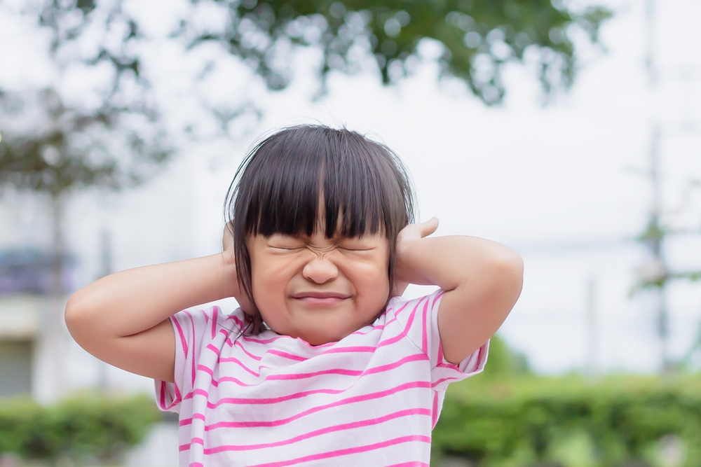 A young asian girl wearing a pink and white striped T-shirt covering her ears and making a grimace.