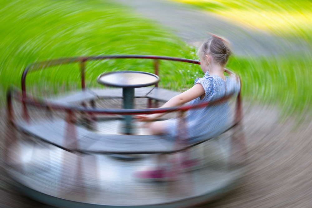 A young blond-haired girl in a dress sitting and spinning on a merry-go-round.
