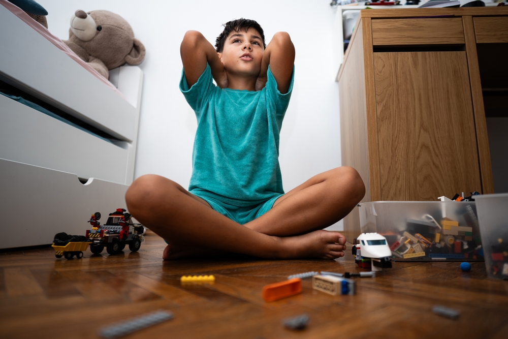 A kid dressed in a green T-shirt and shorts, covering his ears while sitting on his bedroom floor playing with his toys.