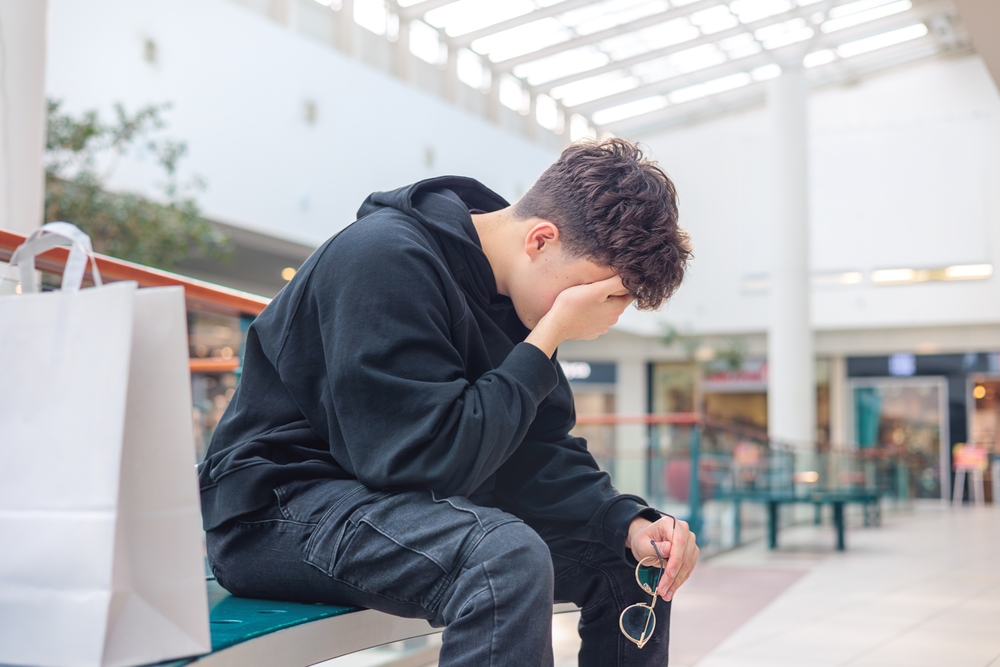 A teenager dressed in a black hoodie and jeans resting his head in his hands at the mall.